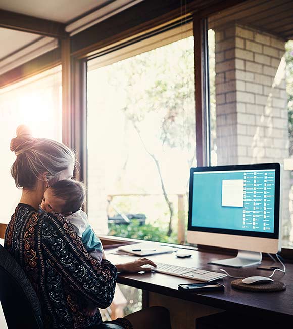 mother holding her newborn baby while working at a computer in her home office