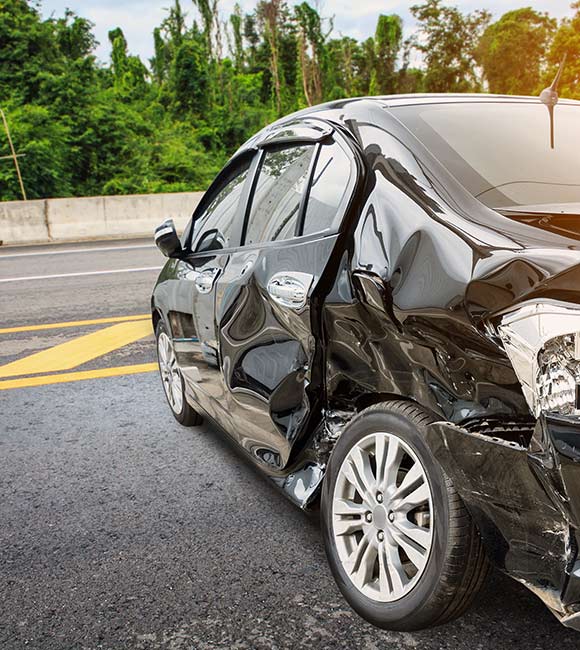 black car sits on the side of a highway with damage to the driver's rear door and tire area